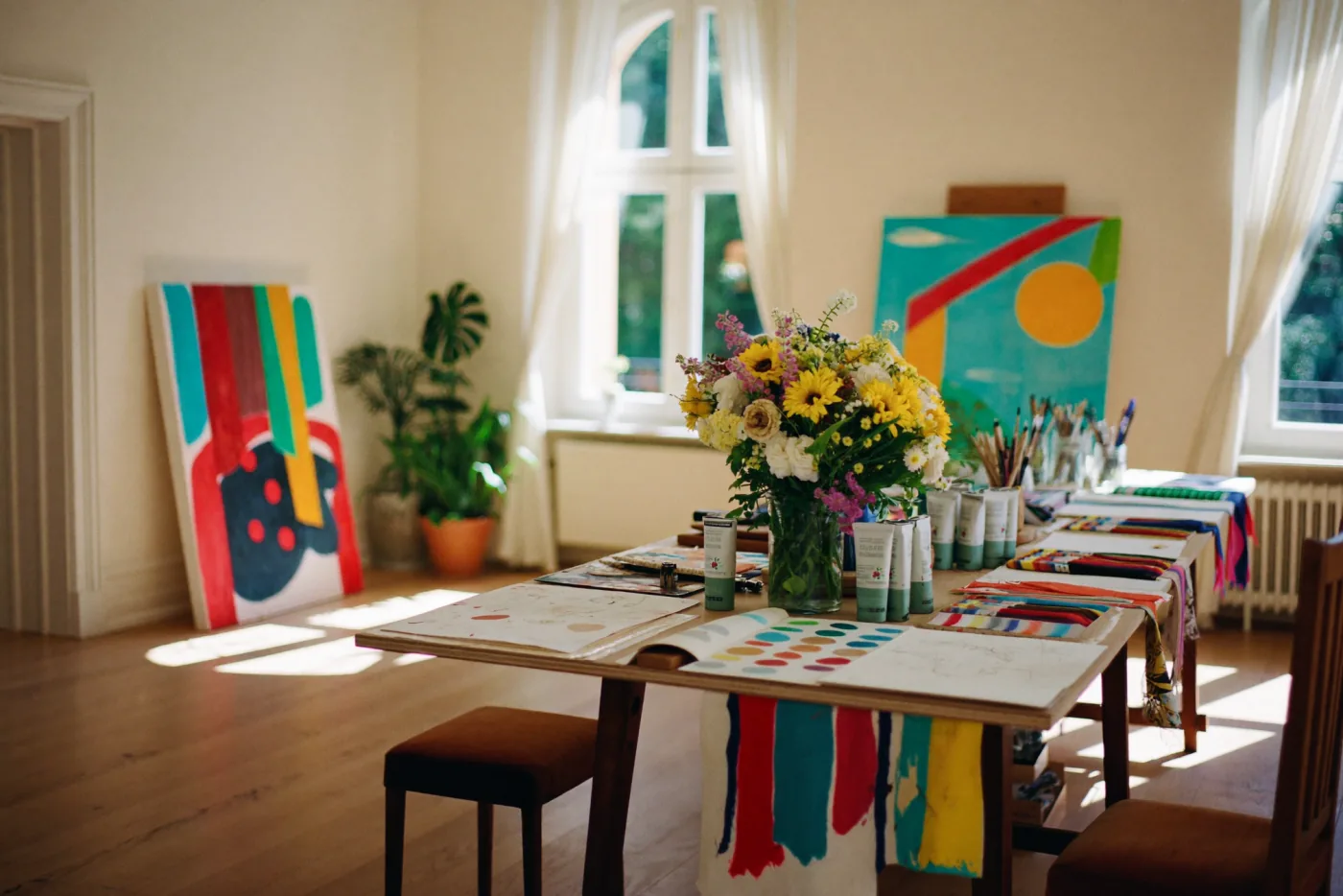 A bright, sun-filled art and design atelier: colourful canvases, painted fabric and flowers on a long worktable