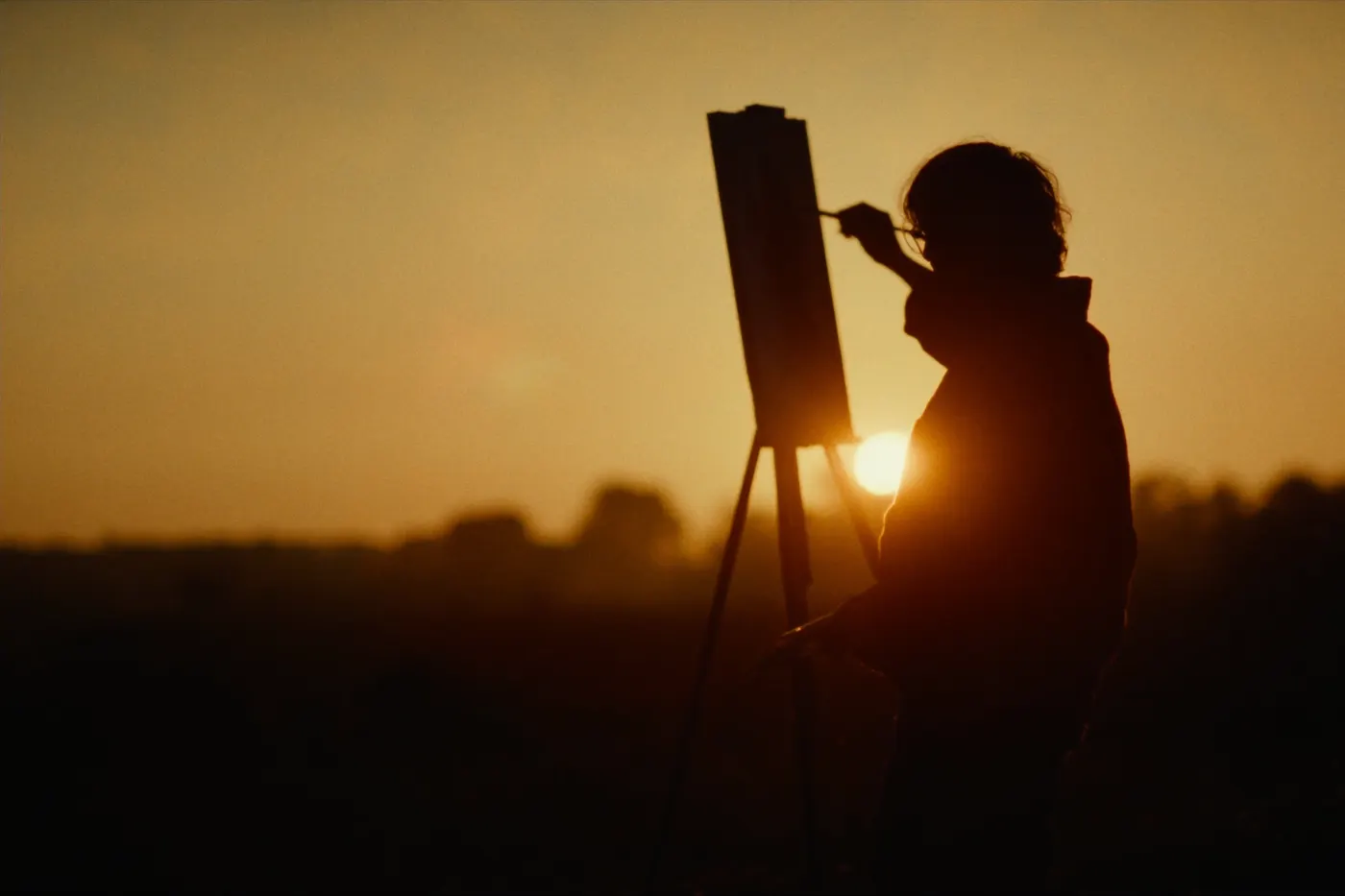 A silhouette of a woman painting at an easel against the setting sun
