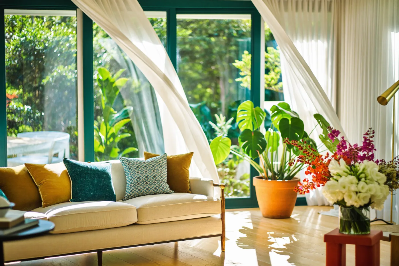 A calm, light-filled living room opening onto lush greenery, with teal and gold cushions and fresh flowers