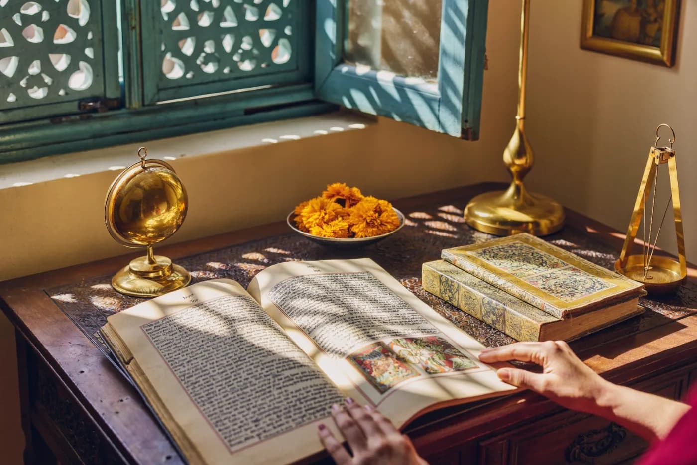 An open Sanskrit manuscript, brass instruments and marigolds on a sunlit desk beside a carved jaali window